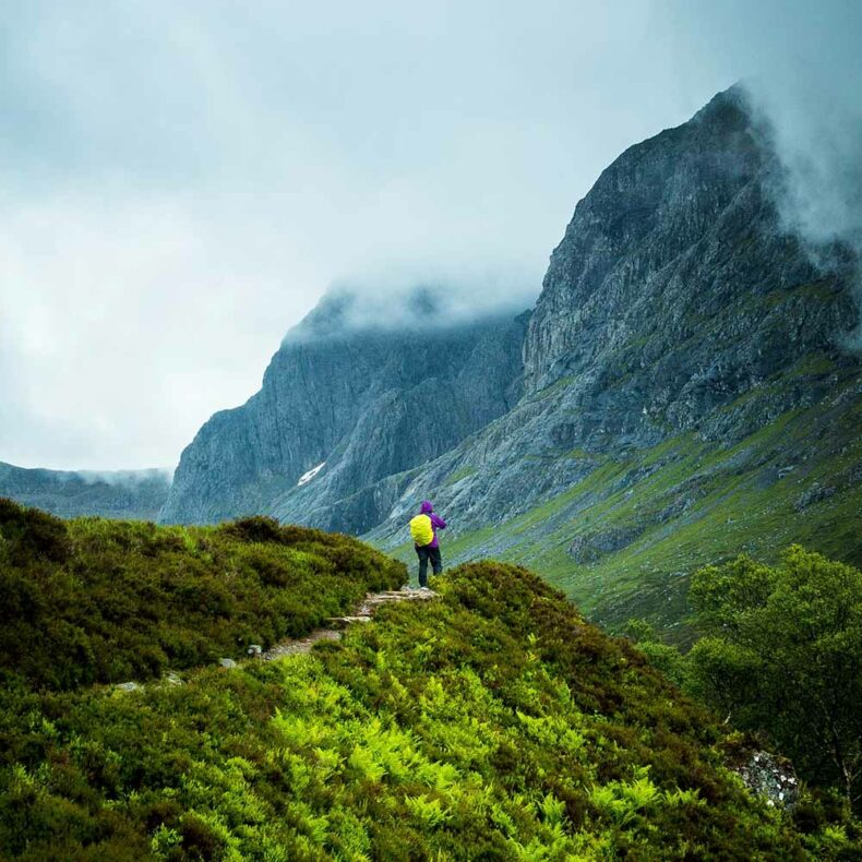 A hiker on a mountain path overlooking the expansive landscape of the Scottish Munros