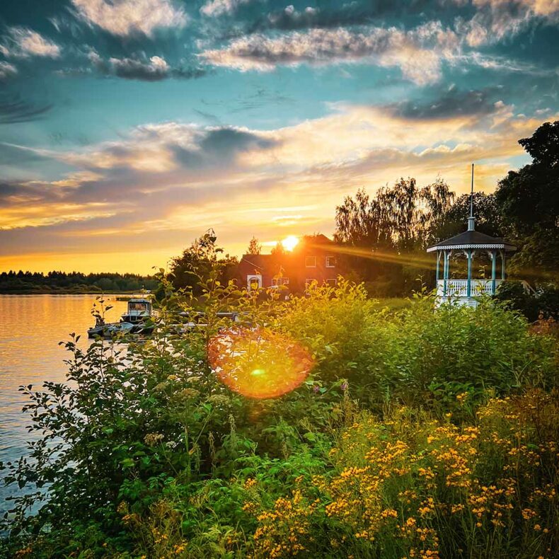 Beautiful sunset at Pikisaari Island, Oulu, Finland, with a red riverside house, white gazebo, boats, and yellow wildflowers