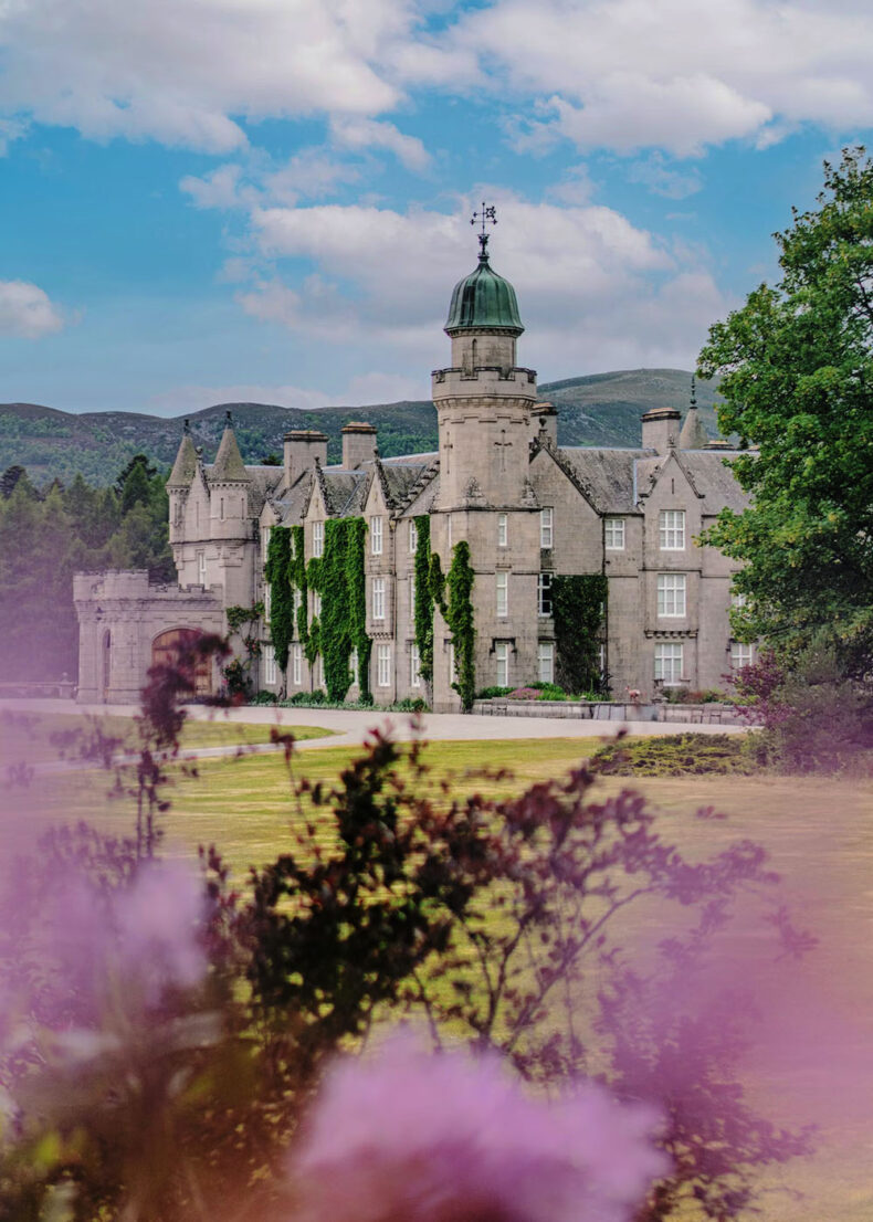 Exterior view of Balmoral Castle in Scotland, featuring its iconic clock tower, grey stone turrets, and lush green gardens in the foreground