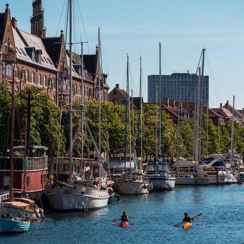 Two people kayaking in a Copenhagen canal lined with docked sailboats and historic brick buildings on a sunny day