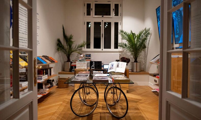 A room at La Mița Biciclista in Bucharest. In the center, a creative display table is constructed from bicycle wheels and a frame, topped with a glass surface. This table and surrounding built-in bookshelves hold numerous art and fashion books