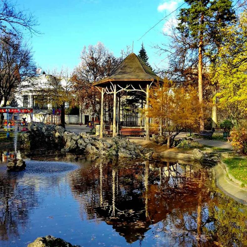 Ioanid Park in Bucharest, featuring a central ornamental pond with a small fountain. A rustic wooden gazebo with a dark roof is centered behind the pond